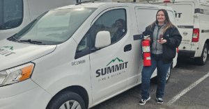 A female Summit Fire & Security technician holding a red fire extinguisher and a tablet, standing next to a white company service van.
