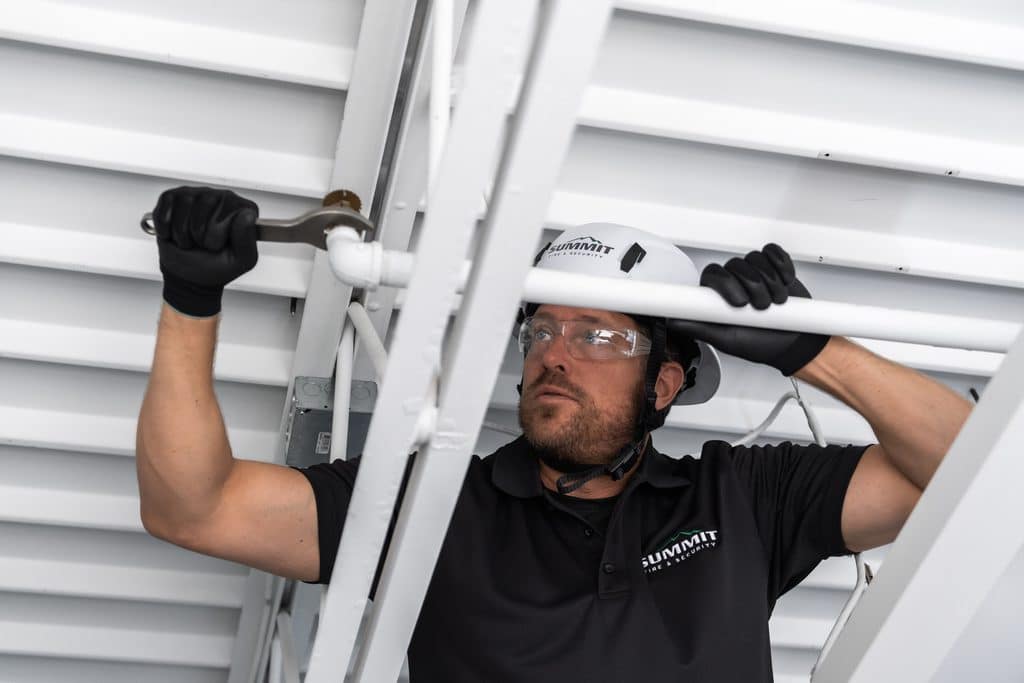 Summit Fire & Security technician wearing a hard hat, safety glasses, and gloves uses a wrench to work on overhead sprinkler piping.
